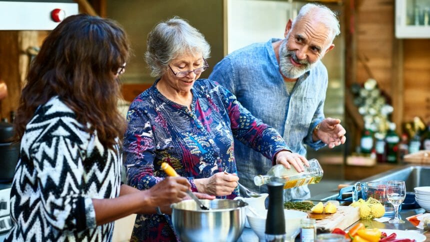 Woman wearing glasses pouring oil and making food, mature man with beard talking to female friend, working as a team, three people cooking at home
