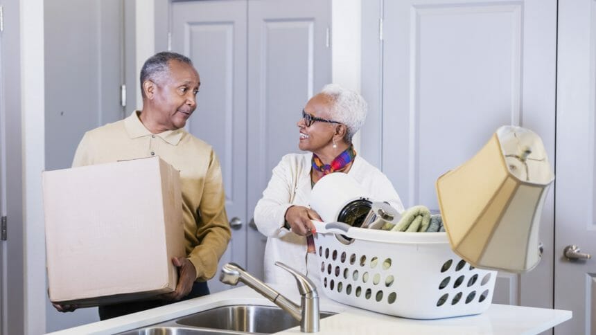 A senior African American couple moving into a new home. They walked through the doorway carrying a box and laundry basket full of personal items and set them on the kitchen counter. They are looking at each other.