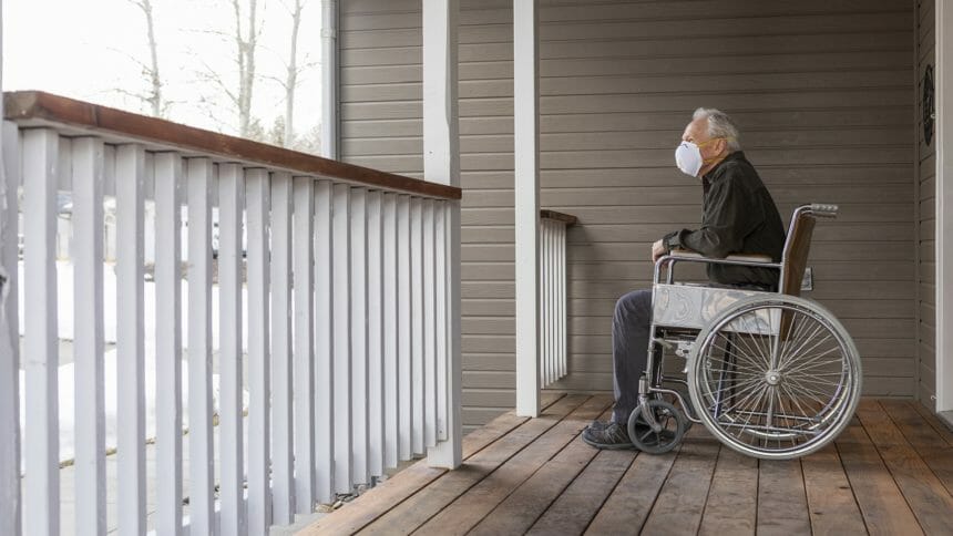 Senior man in wheelchair wearing protective mask to prevent coronavirus transmission on porch