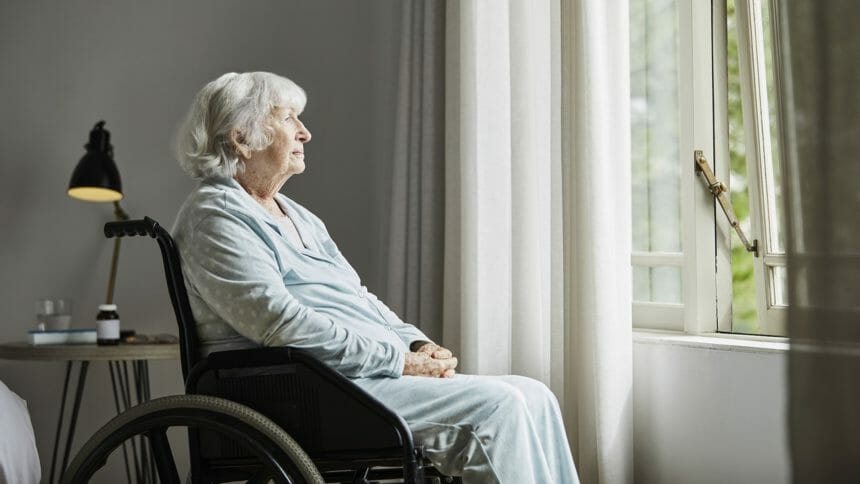 Contemplated senior woman sitting on wheelchair. Elderly disabled female is looking through window.