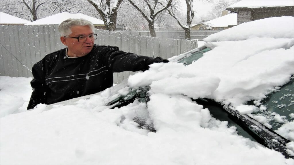 Older man with glasses cleans snow off car in Texas in January 2021