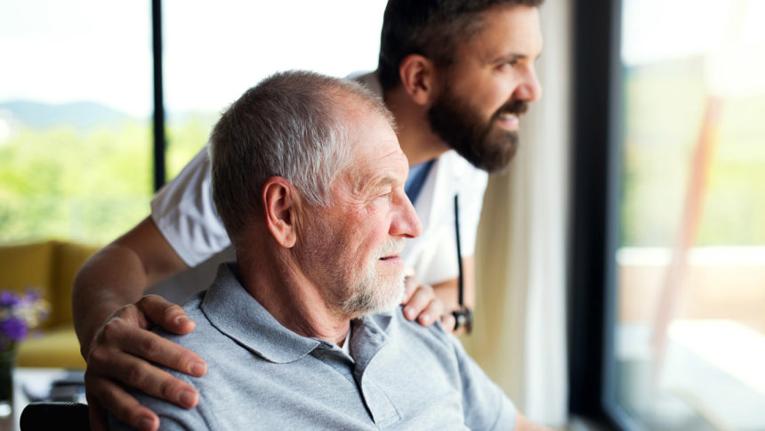 Healthcare worker holding the shoulders of an older man looking out a window
