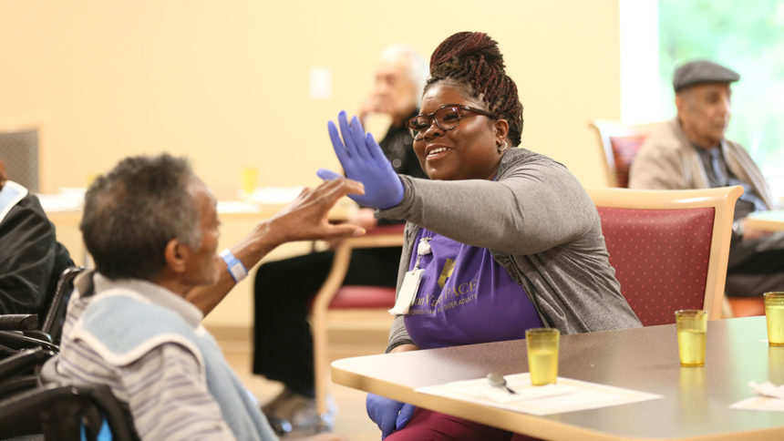 Caregiver in gloves high-fiving a resident sitting at a table.