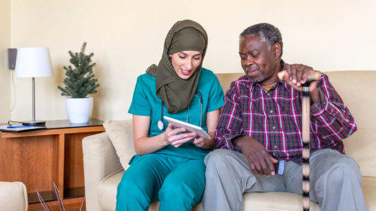 Arabic nurse holding tablet for older adult, sitting on couch.