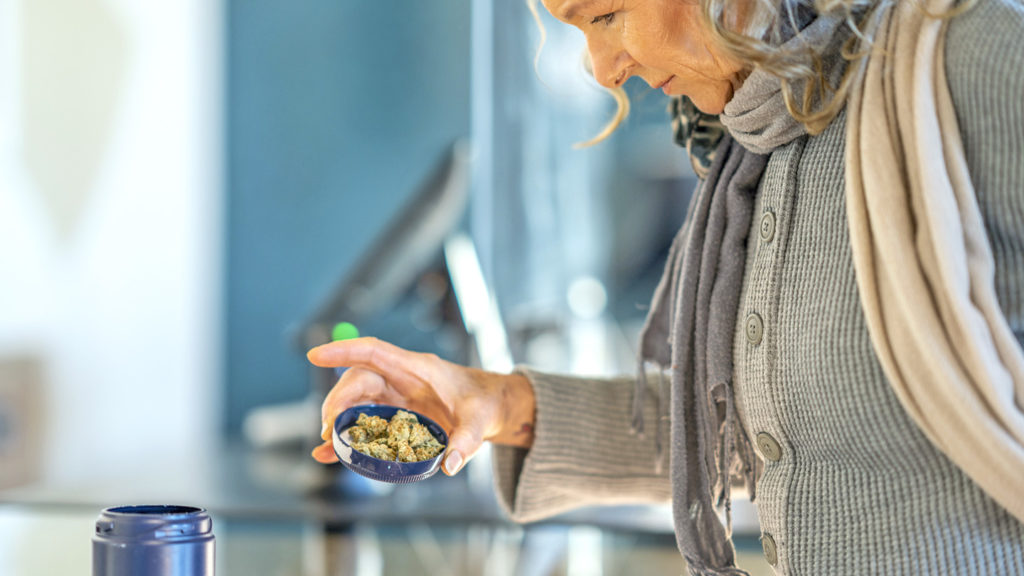 A mature woman leans in over a counter and reaches for some cannabis buds that have been laid out by the associate in the product lid. The woman is inspecting the product before purchasing from the legal retailer.
