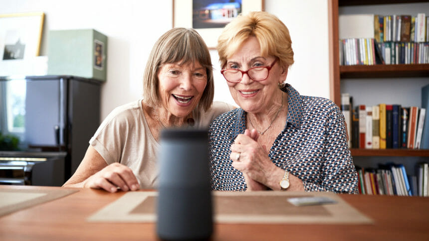 Two senior women looking excited while taking with an interactive voice assistant smart speaker. Excited elderly female friends asking questions to a digital assistant at home.
