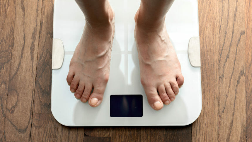 Top down view of feet standing on white digital bathroom scale over wooden floor. Dieting, healthy lifestyle and achieving weight goals concept.