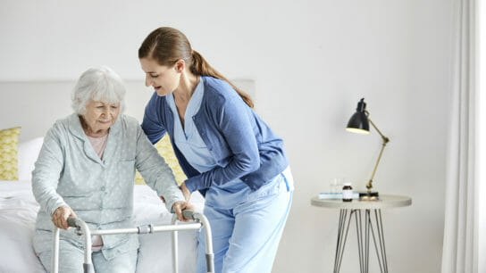 Nurse supporting senior woman with mobility walker to stand. Caregiver is assisting disabled female in recovery. They are in bedroom at home.