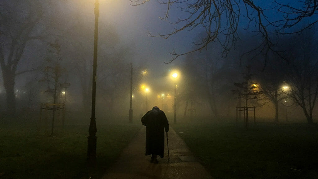 Rear View Of Senior Person Walking On Footpath In Park At Night During Foggy Weather