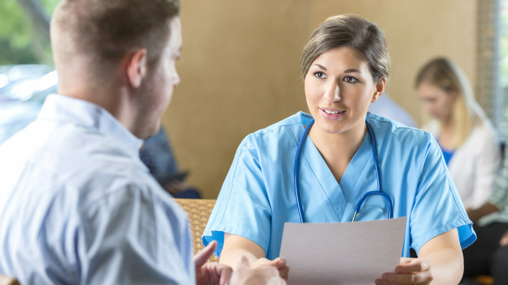 Mid adult Caucasian woman is manager of nursing staff at hospital. She is reading a resume and interviewing a male candidate for a healthcare job. Other potential employees are waiting to be interviewed during hospital's staff recruitment event.