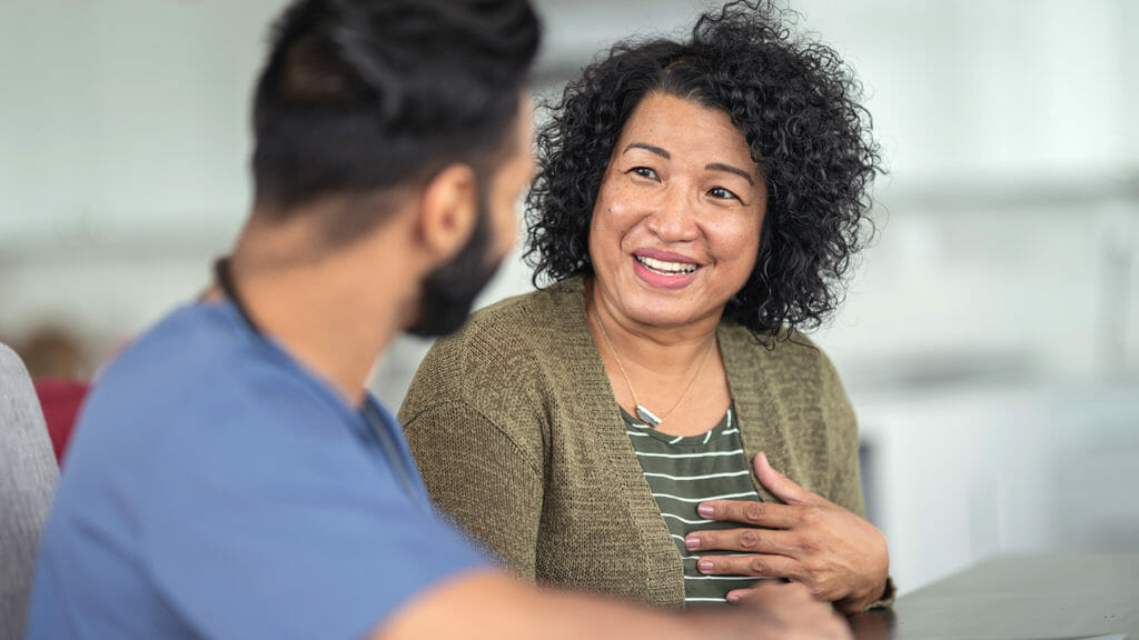 A woman of Asian descent is at a medical consultation. She is sitting at a table next to her doctor. The doctor is a mixed-race man of Asian and Indian descent. The two individuals are having a conversation. The patient is smiling and talking.