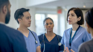 A small group of diverse medical staff stand around together in the hallway of a hospital as they have a brief meeting to discuss a patient case. They are each wearing scrubs and name badges as they focus on the conversation.