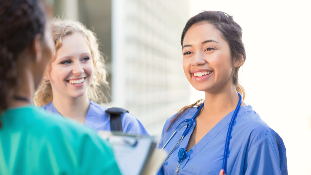 Young adult Hispanic woman is talking with young adult Caucasian blonde woman and young adult African American woman outside on college campus. Women are nursing or medical students. They are wearing hospital scrubs and stethoscopes.