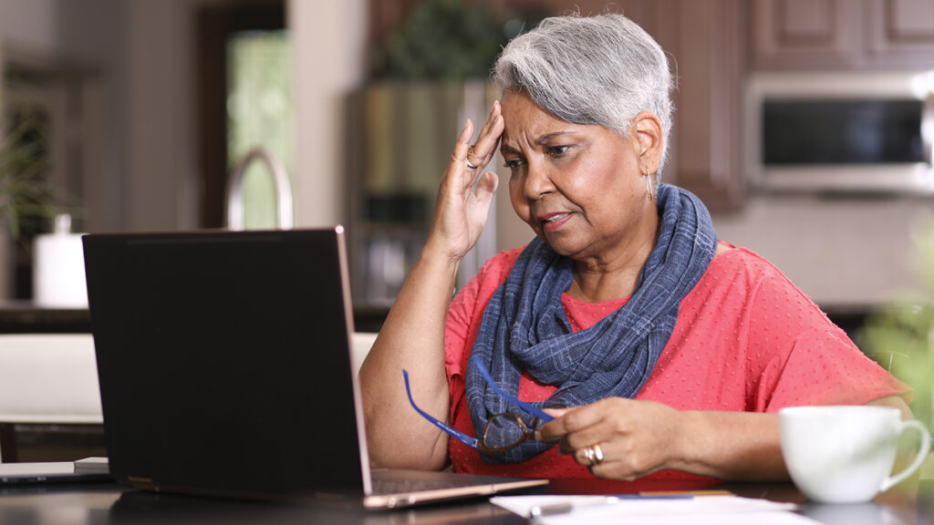 African descent, senior adult woman at home sitting at kitchen table paying bills or online banking using laptop computer. She is frustrated at the overdue charges. Paperwork and coffee cup on table.