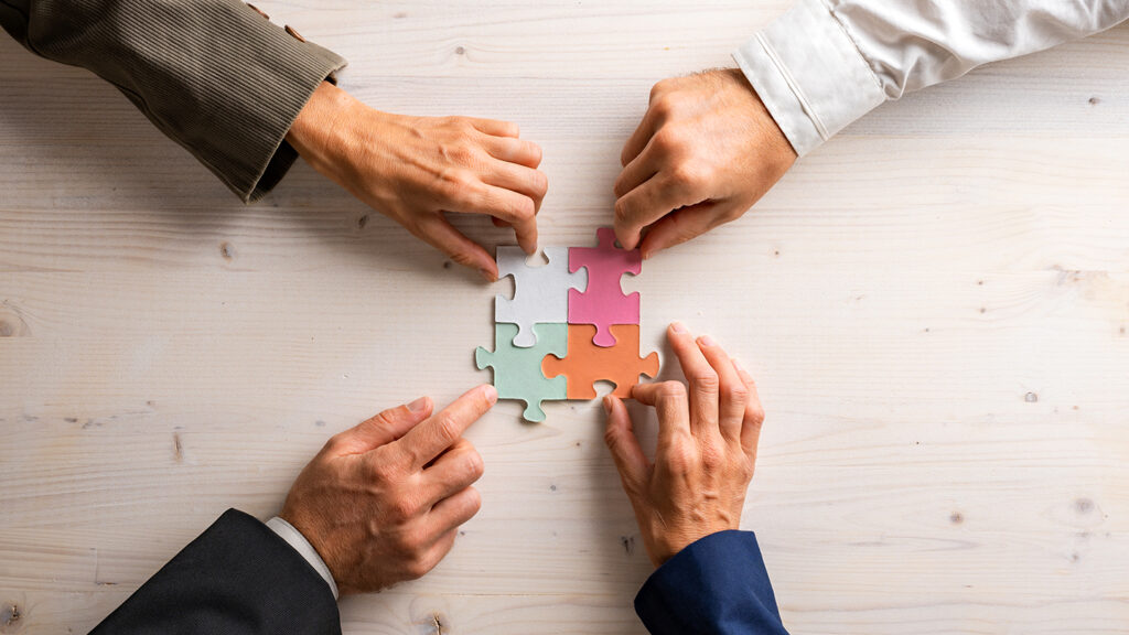 Hands of four businesspeople joining matching puzzle pieces of various pastel colors over a wooden background.
