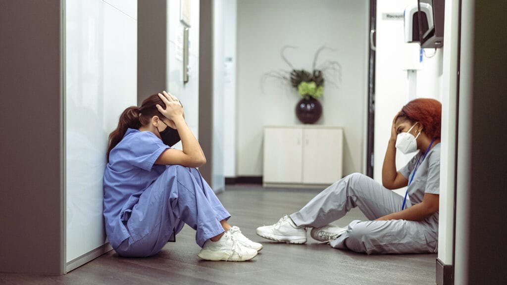 Two nurses appear stressed and tired. They are seated on the hospital hallway floor. They are comforting one another after a stressful day attending to patients with covid-19. They welcome the break from the chaos of the hospital wards.