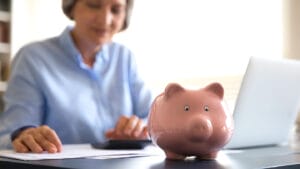 Classic piggy bank on desk, older woman sit at workplace with laptop use calculator counts monthly payments, on background. Taxes, incomes calculation, finances management, save money, savings concept
