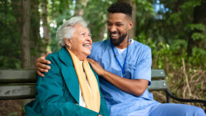 Happy caregiver man helping and supporting senior woman sitting outdoors in park.