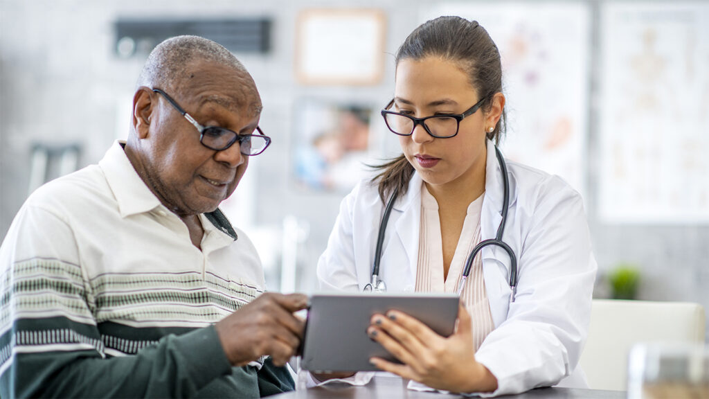 A senior black man is sitting in his doctor's office and listening as the doctor shows him something on a digital tablet.