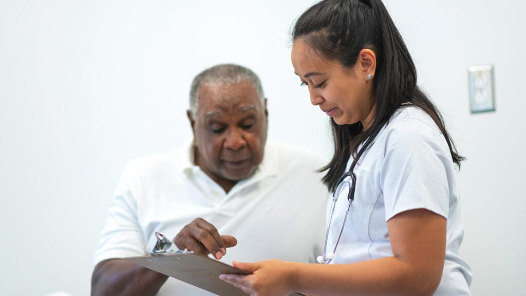 A female doctor of Asian descent is consulting a black senior male patient. The patient and doctor are sitting next to each other in a medical clinic examination room. The doctor is showing the patient test results on a clipboard.